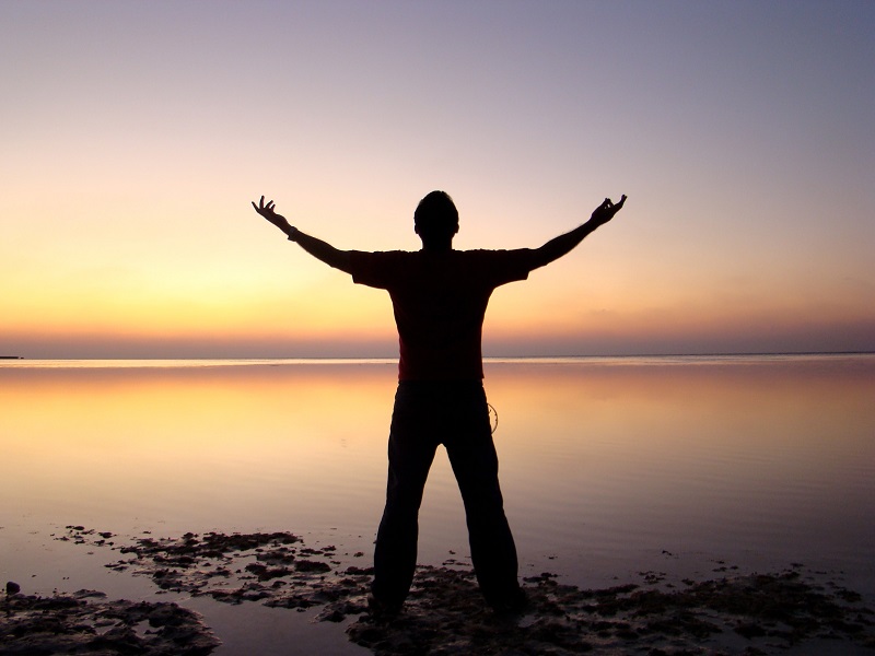 Silhouette of man standing in front of water at dusk with arms outstretched