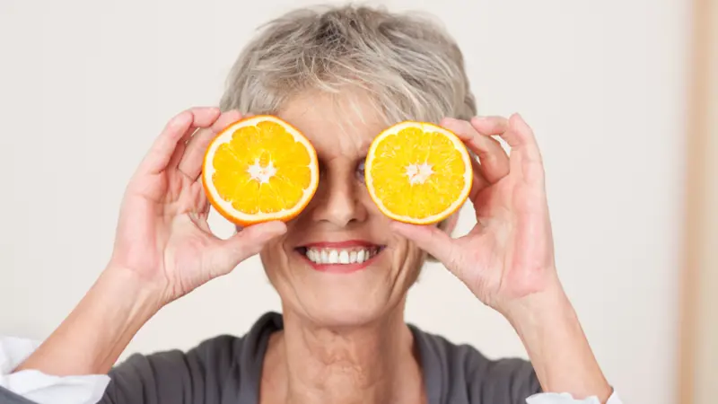 Smiling senior woman holding half oranges over eyes against white background