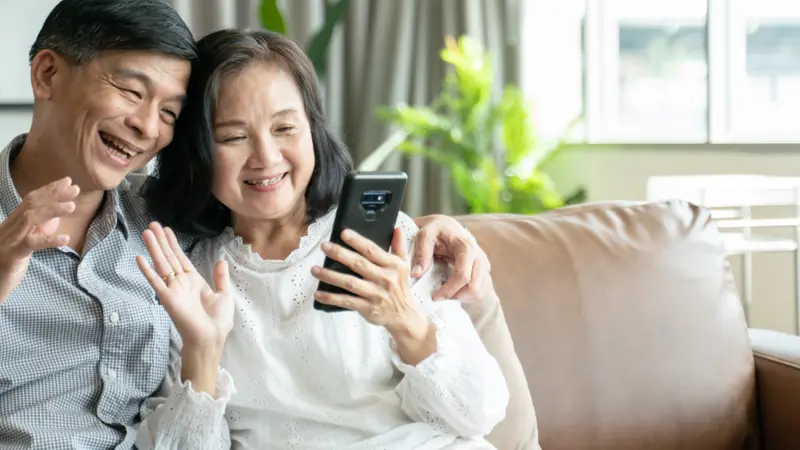 couple sitting on sofa in home playing smartphone and video conference with grandchild