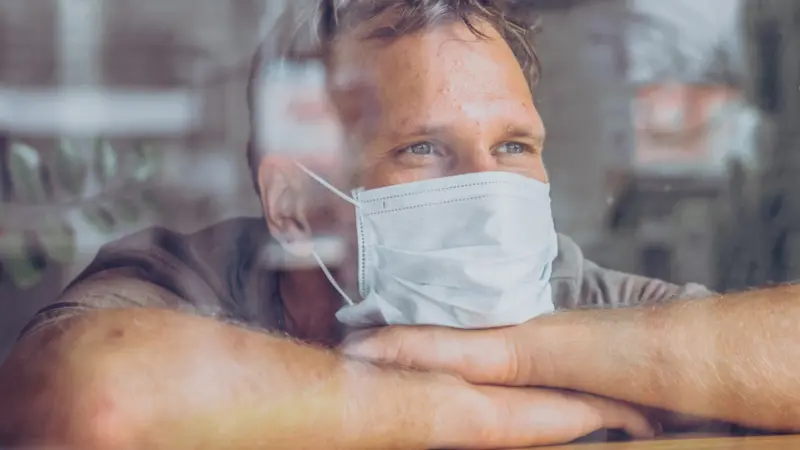Man wear white protective mask, looking outside through window, self isolation due to global COVID 19 Coronavirus pandemic