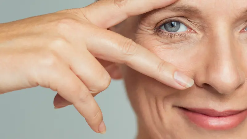 smiling senior woman showing victory sign near eye on grey background