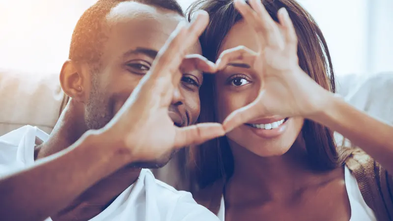 couple sitting close to each other and looking through a heart shape made with their fingers couple sitting close to each other and looking through a heart shape made with their fingers
