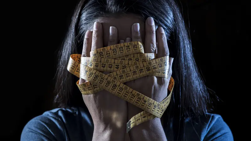 close up hands wrapped in tape measure covering face of young girl suffering nutrition disorder on black background