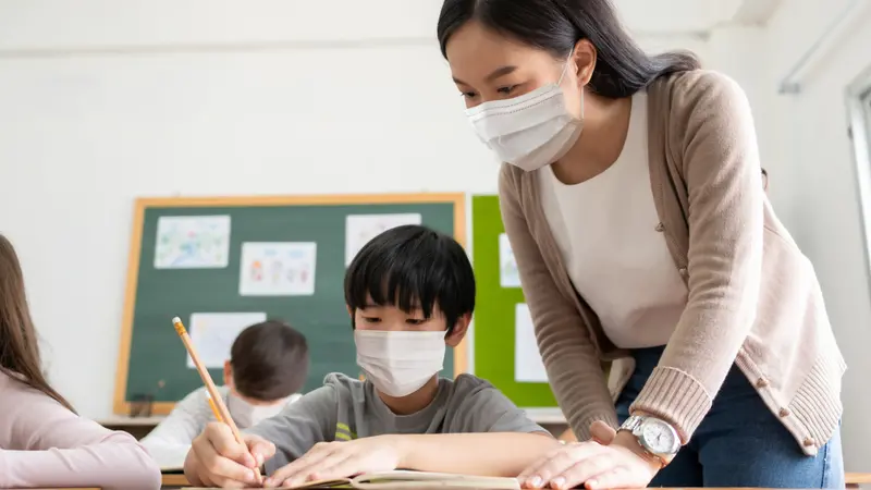 Teacher with little boy wearing protective face masks studying in classroom during COVID-19 pandemic