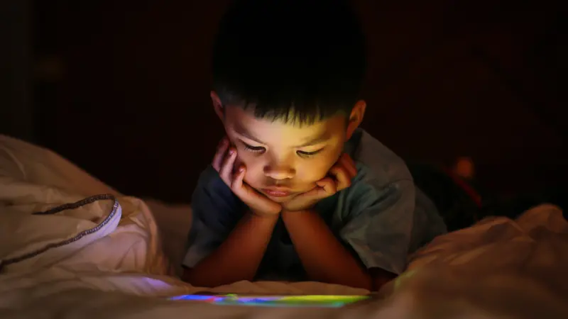 kid alone watching tablet device, lying on white duvet bed with chin on hands, in background darkness bedroom night time.