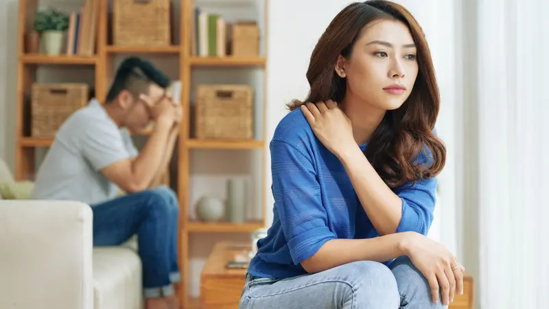 Sad Asian woman sitting alone with man on background being in conflict at home