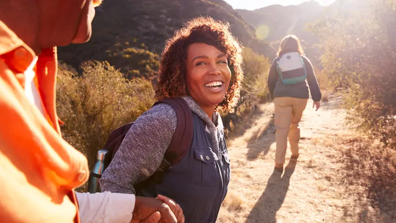 Woman Helping Man On Trail As Group Of Senior Friends Go Hiking In Countryside Together