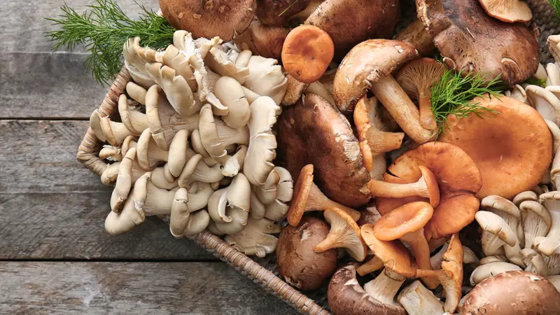 Wicker tray with variety of raw mushrooms on wooden table