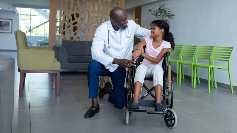 doctor kneeling to talk with girl in wheelchair in hospital foyer. medicine, health and healthcare services.