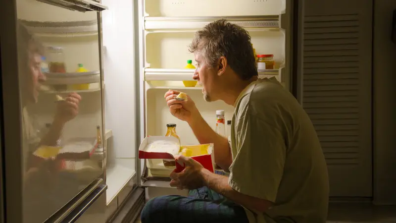 A man with sleep-related eating disorder sleep sits in front of a refrigerator eating out of carton