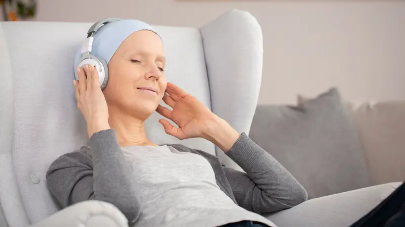 Peaceful woman with headphones and blue headscarf resting at home after cancer treatment