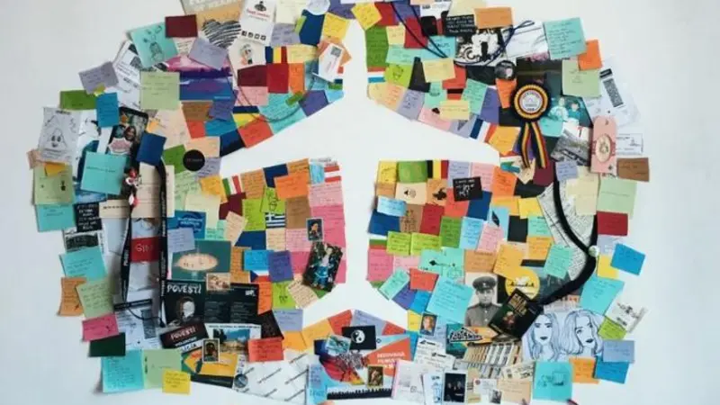 White wall with scraps of paper, pictures and post it notes in a circle with a vacancy in the shape of an airplane in the middle. A hand reaches out towards the wall at the bottom. 