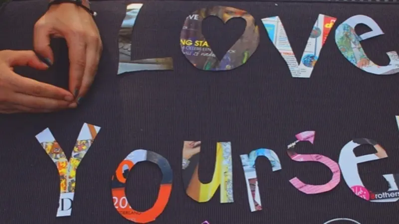 Feel the Love Hands making a heart shape with collage letters reading love yourself on a black background