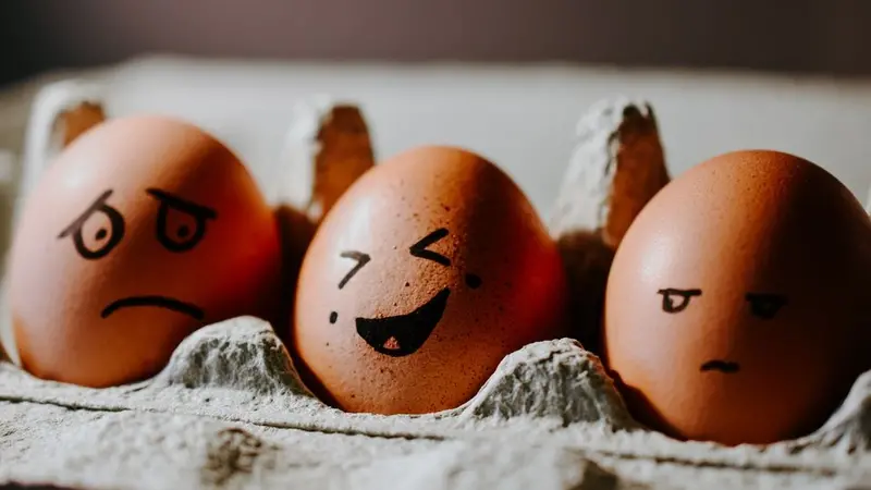 Three brown eggs in an egg carton with black sharpy emotional faces drawn on them, sad, laughing and glaring