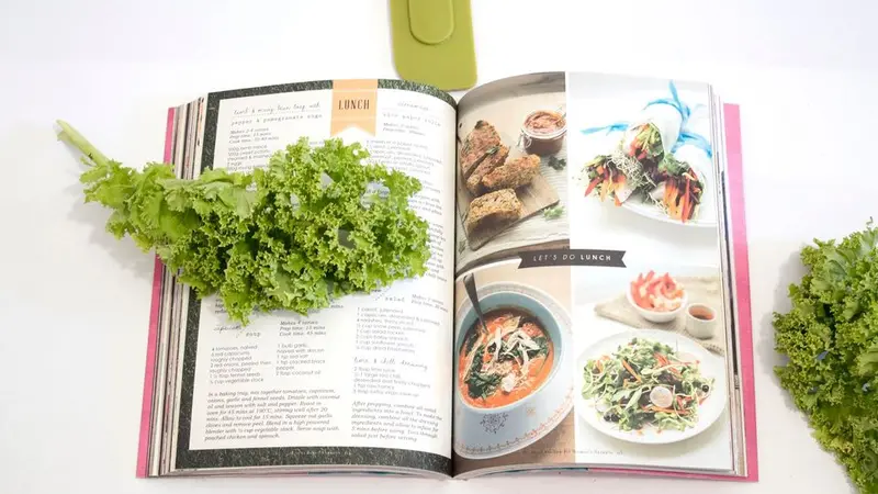 Cookbook lying open on white table with a piece of kale draped over the left page