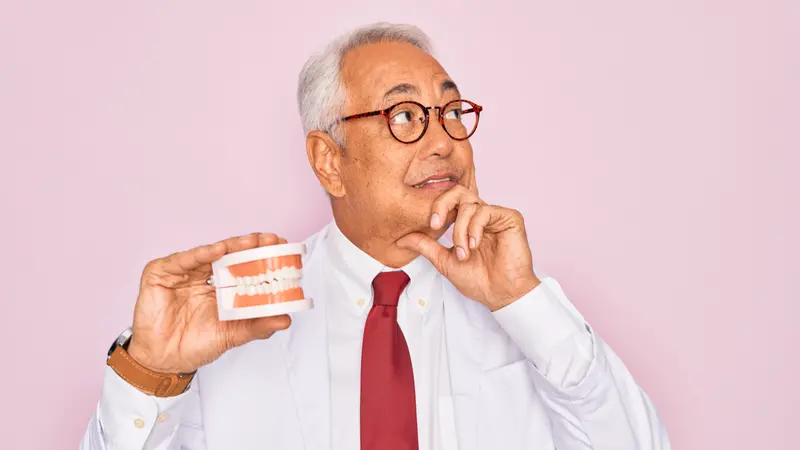 Middle age senior grey-haired dentist man holding prosthesis denture over pink background, confused 