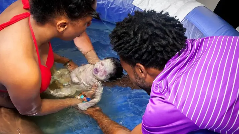 Woman in a red bikini in a blue birthing pool with a newborn baby, a man in a purple stripped shirt is leaning into the pool and supporting the baby's body with his hands