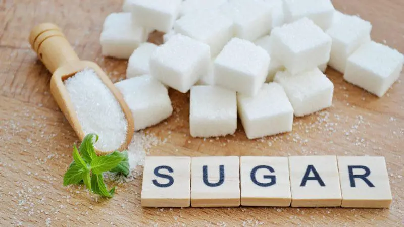 wooden table with wooden scoop full of sugar surrounded by a pile of sugar cubes and wooden scrabble tiles spelling out the word sugar