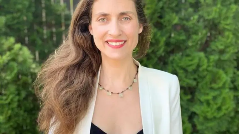 image of a woman, Dr. Liza Cherubinia, long brown hair, white suit jacket standing outside in front of evergreens