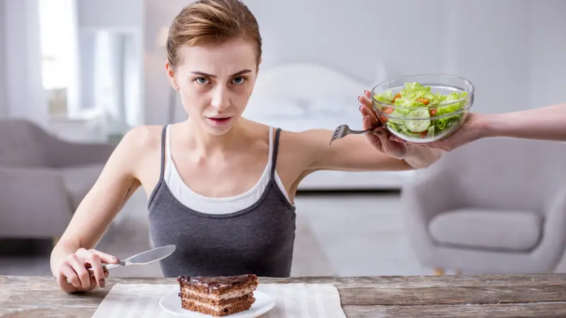 Depressed young woman refusing from the salad while wanting to eat the cake