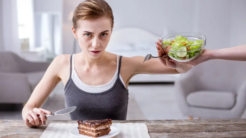 Depressed young woman refusing from the salad while wanting to eat the cake Depressed young woman refusing from the salad while wanting to eat the cake