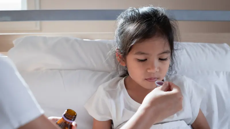 Asian mother giving cough syrup medicine on a spoon to her daughter in bed Asian mother giving cough syrup medicine on a spoon to her daughter in bed