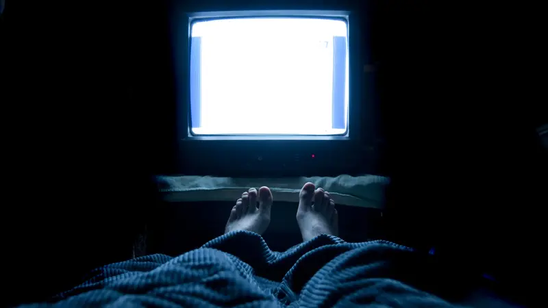 Person sleeps in his bed with TV on at night with his feet sticking up out of the blankets. 
