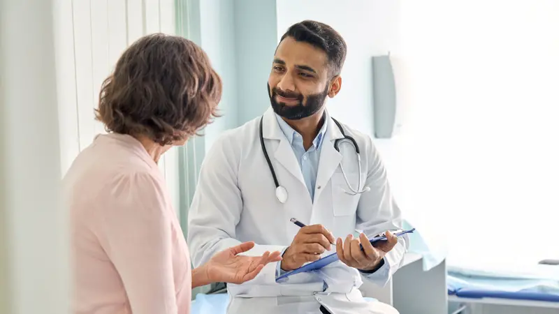 Indian male doctor consulting senior old patient filling form at consultation.