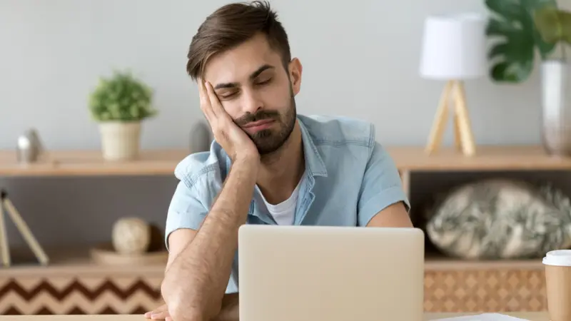 Exhausted millennial male hold head with hand sitting at office table falling asleep