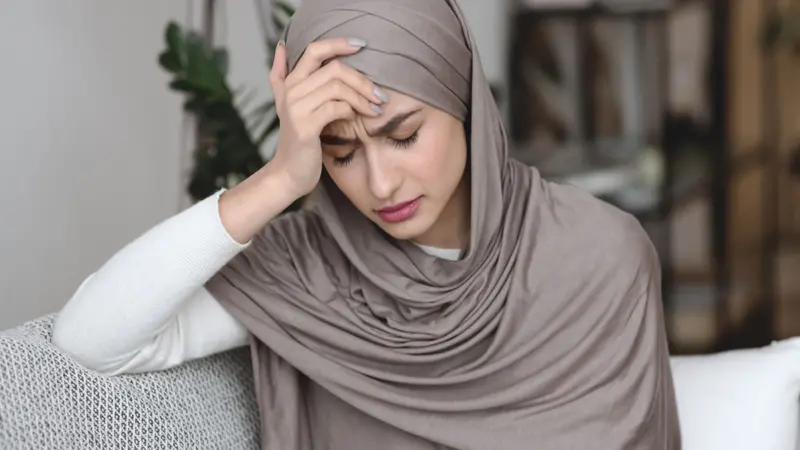 Muslim woman in headscarf sitting on sofa and touching head with headache