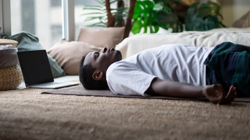 African young man meditating on a floor at home using Yoga nidra practice online.