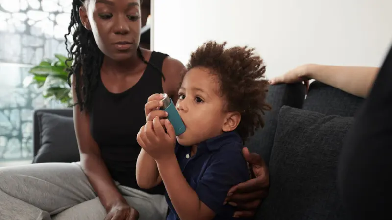 Worried mother looking at son using inhaler when having asthma attack at home