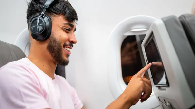 Latin American man in plane cabin using smart device listening to music on headphones. Latin American man in plane cabin using smart device listening to music on headphones.