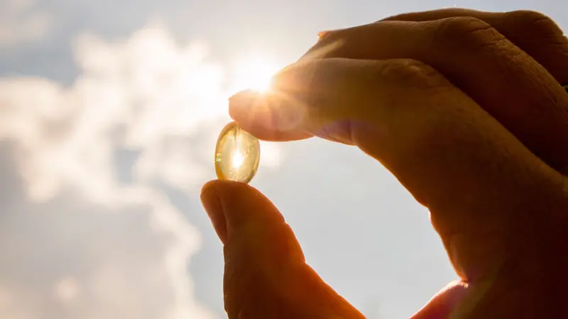 Yellow soft shell D-vitamin capsule against sun and blue sky on sunny day.