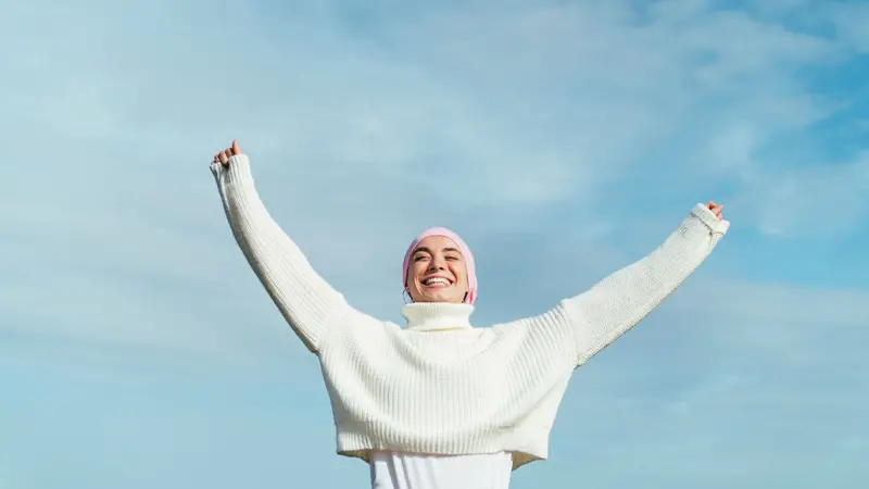 Happy young woman with cancer with arms up. She is wearing a pink scarf on her head and white jersey.