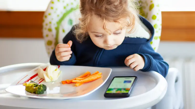 Toddler eats in the high chair while watching movies on the mobile phone.