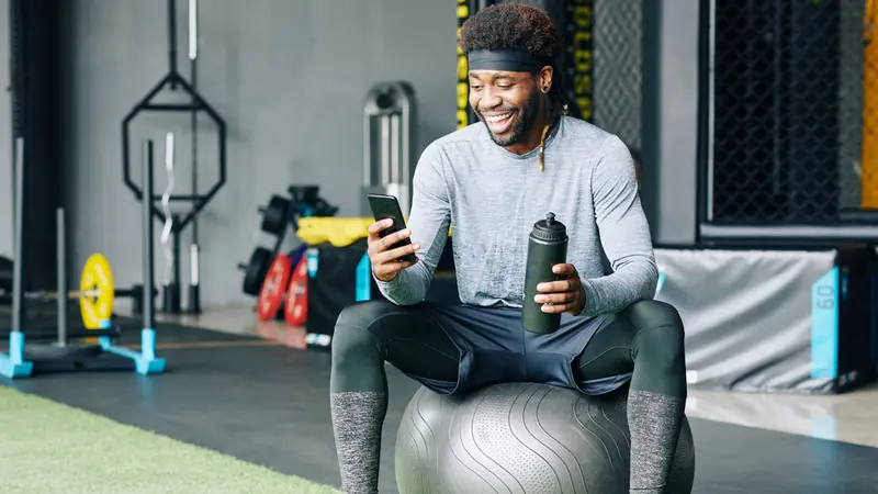 Excited young Black man sitting on fitness ball with bottle of water and checking social media on smartphone