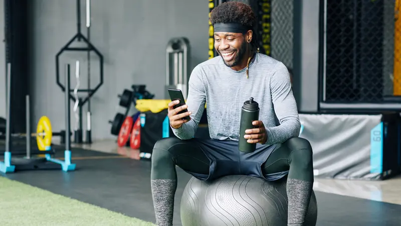 Excited young Black man sitting on fitness ball with bottle of water and checking social media on smartphone Excited young Black man sitting on fitness ball with bottle of water and checking social media on smartphone