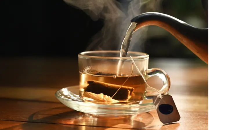 Hot water pouring into transparent cup with tea bag inside on wood table in morning sunlight