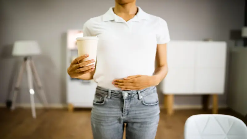 Person holding a cup of coffee with one hand and their stomach with another hand Person holding a cup of coffee with one hand and their stomach with another hand