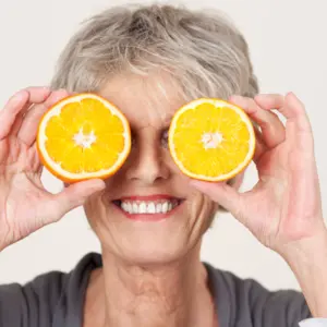 Smiling senior woman holding half oranges over eyes against white background