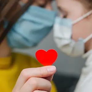 mommy hugs daughter in protective medical masks sitting on bed, holding red heart