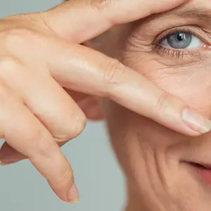 smiling senior woman showing victory sign near eye on grey background