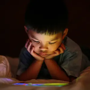 kid alone watching tablet device, lying on white duvet bed with chin on hands, in background darkness bedroom night time.