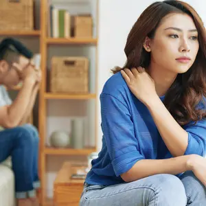 Sad Asian woman sitting alone with man on background being in conflict at home