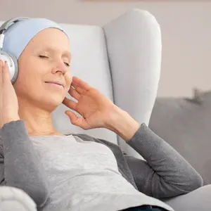 Peaceful woman with headphones and blue headscarf resting at home after cancer treatment