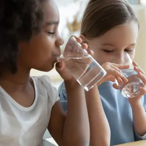Two multi racial little girls sit at table in kitchen drinking clean water close up image.