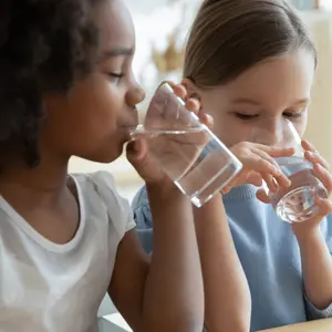 Two multi racial little girls sit at table in kitchen drinking clean water close up image. Two multi racial little girls sit at table in kitchen drinking clean water close up image.