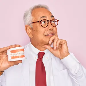 Middle age senior grey-haired dentist man holding prosthesis denture over pink background, confused 