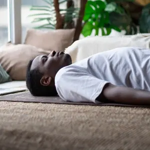 African young man meditating on a floor at home using Yoga nidra practice online.